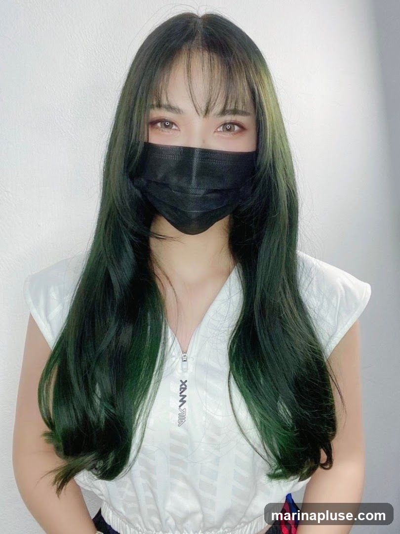 A woman with long, dark green hair and bangs is wearing a black face mask and a white sleeveless top with a zipper. The top has a subtle pattern and a logo on the front. She is looking directly at the camera. The background is a plain white wall.