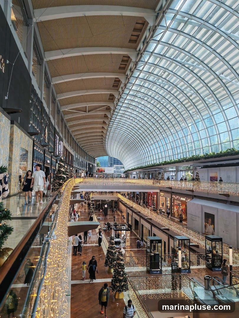 The image shows the interior of a large, multi-level shopping mall. The architecture features a curved glass ceiling and wooden paneling. Numerous shops are visible on different levels, with escalators and walkways connecting them. The mall is decorated for Christmas with garlands of lights along the railings and several Christmas trees. Many shoppers are present, browsing the stores and walking around.