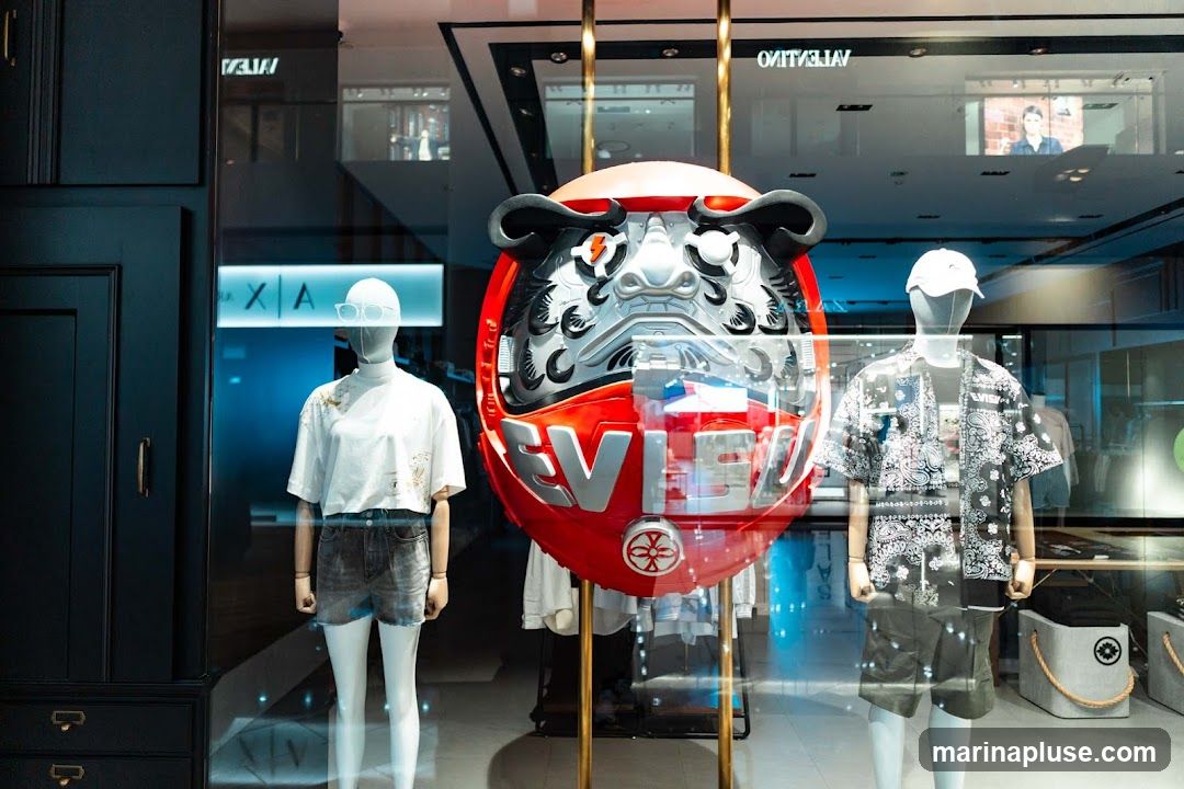 A store window display features two mannequins, one on the left wearing a white t-shirt and denim shorts, and another on the right wearing a black bandana-print shirt, t-shirt, and shorts. In the center, a large, red, robotic-looking Daruma doll with the brand name "EVISU" is prominently displayed. The reflection in the window shows the interior of the store, including other displays and signage, as well as the exterior environment.
