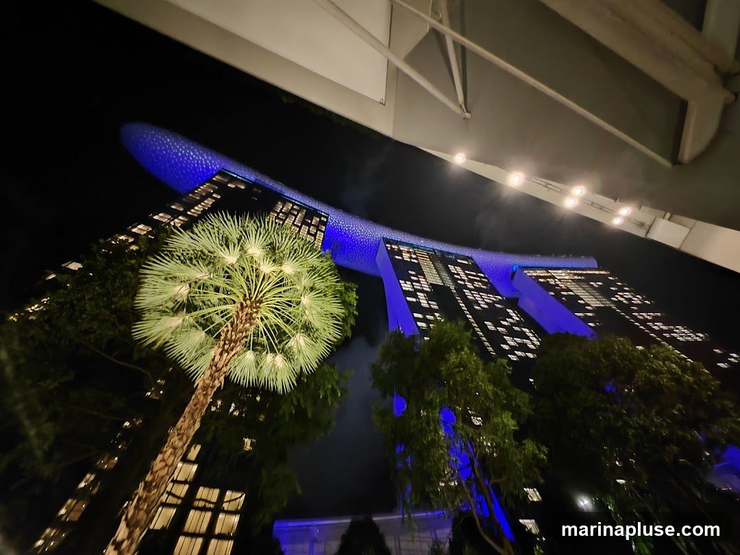 A low-angle night view of the Marina Bay Sands hotel in Singapore. The iconic building is illuminated with blue lights, and its windows are lit up. A palm tree and other greenery are in the foreground, with a walkway structure overhead.