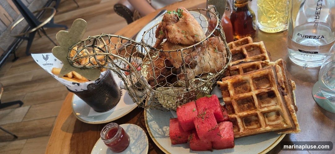 A close-up shot of a meal featuring fried chicken served in a wire basket shaped like a chicken, accompanied by waffles, watermelon cubes, and french fries in a paper-lined bucket. A small jar of red sauce, a bottle of syrup, and a bottle of Purezza sparkling water are also visible on the table.