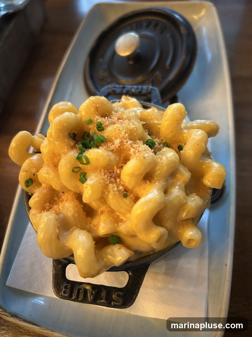 A close-up shot of macaroni and cheese served in a small black Staub pot. The pasta is coated in a creamy yellow cheese sauce and topped with breadcrumbs and chopped chives. The pot is placed on a light blue rectangular dish, which is on a white napkin.