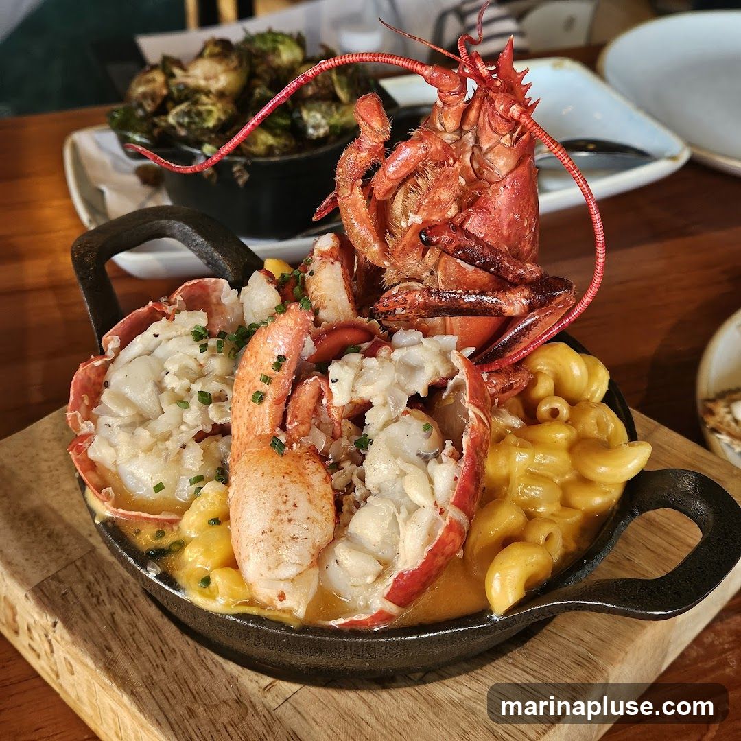 A whole lobster is served atop a dish of macaroni and cheese. The lobster is split lengthwise, revealing its white meat. The mac and cheese is a creamy, golden yellow. In the background, a bowl of roasted Brussels sprouts is visible.