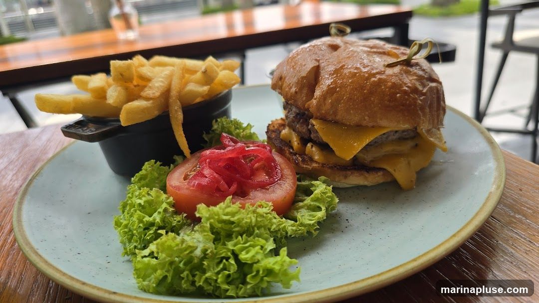 A close-up shot of a cheeseburger served with a side of french fries and a small salad garnish. The burger has two patties and melted cheese, served on a bun. The fries are golden brown and in a small black dish. The salad consists of lettuce and sliced tomatoes topped with red onions. The meal is presented on a light blue plate with a textured rim, placed on a wooden table.