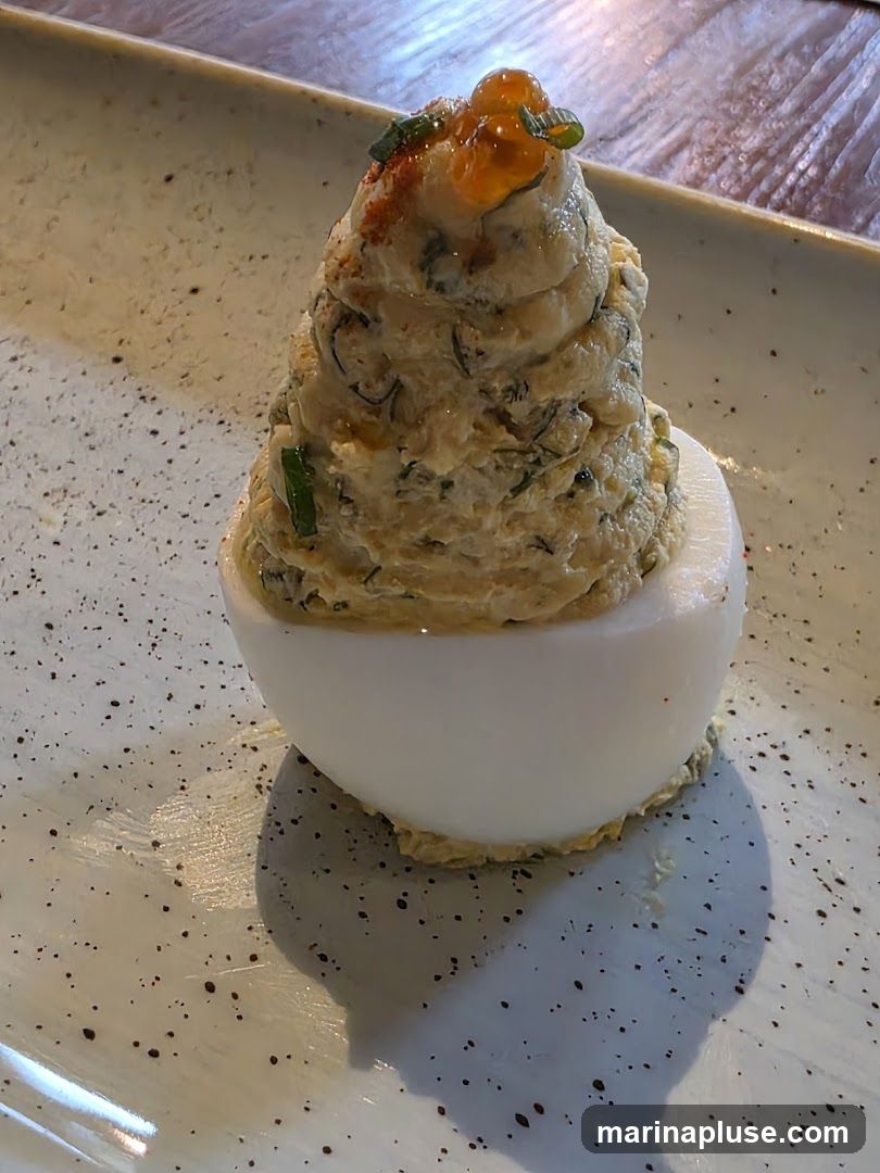 A close-up shot of a deviled egg, topped with a generous swirl of creamy filling, garnished with bright orange caviar and chopped green onions. The egg is served on a speckled white plate, with a wooden surface visible in the background.