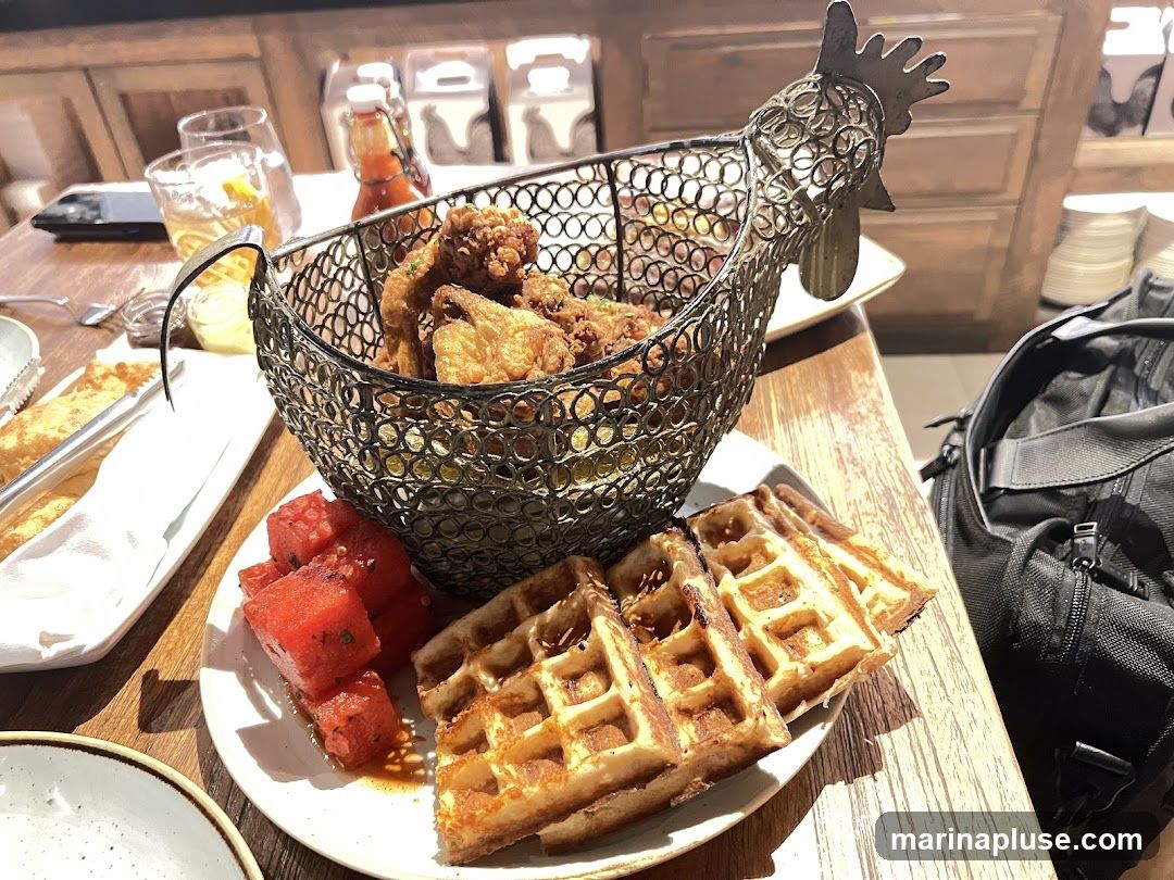 A metal basket shaped like a chicken holds fried chicken pieces. Next to the basket are several waffles and a plate of cubed watermelon. A drink with ice and a lemon slice is visible in the background, along with bottles of sauce. A black bag is on the right side of the table.