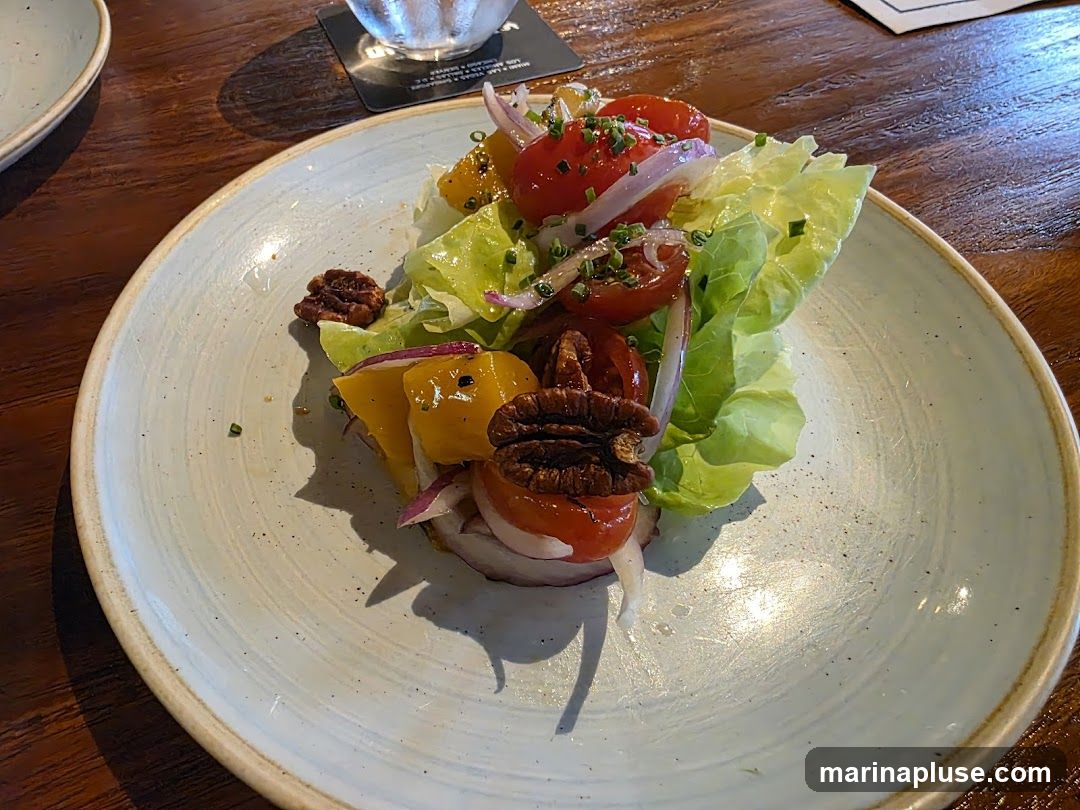 A close-up shot of a salad served on a light blue plate. The salad features lettuce, cherry tomatoes, red onion slices, and pecans. There are also pieces of yellow bell pepper visible. The salad is garnished with chopped chives. The plate is set on a wooden table.