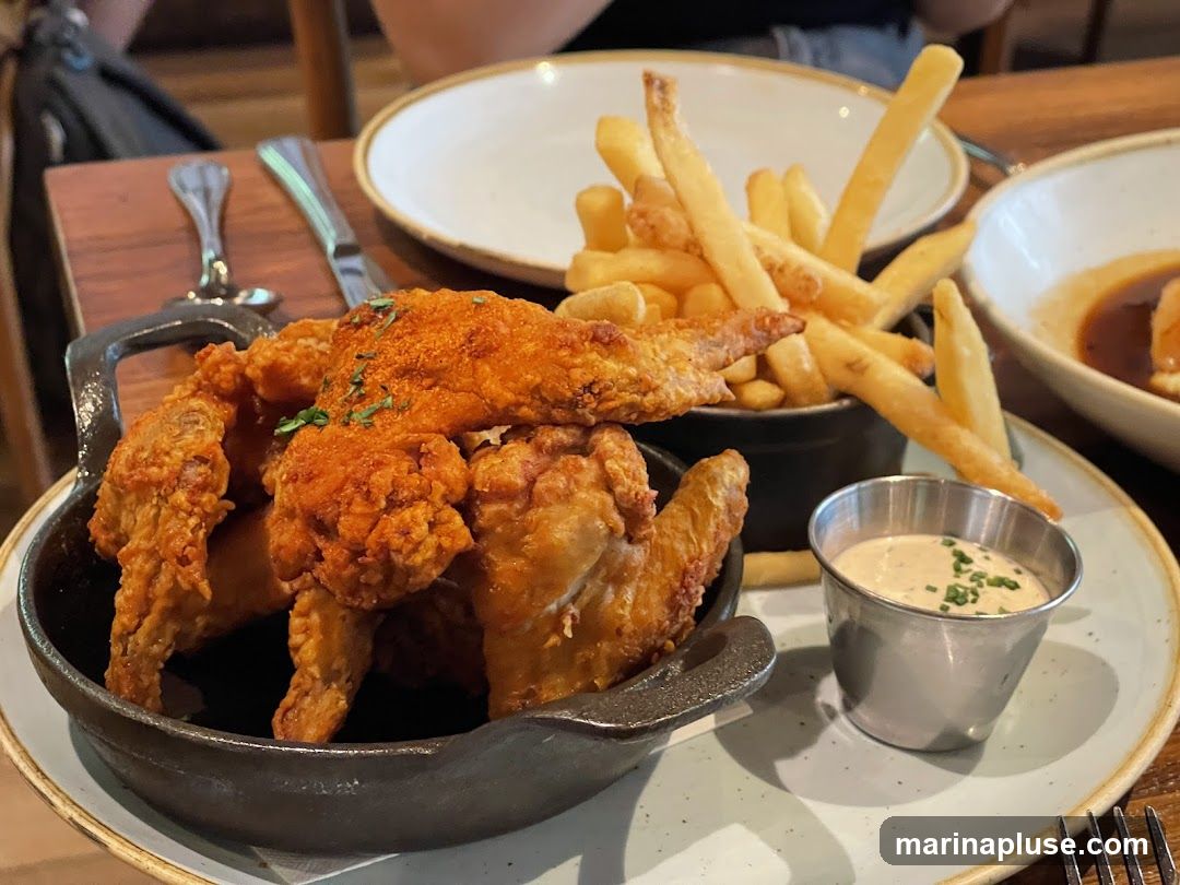 A close-up shot of a serving of fried chicken wings and french fries, presented in a rustic setting. The chicken wings are golden brown and appear to be seasoned with spices. They are served in a dark, cast-iron-style dish. A small metal cup filled with a creamy dipping sauce, garnished with chives, sits next to the wings. A generous portion of french fries is piled in a dark container behind the chicken. In the background, out-of-focus plates and cutlery suggest a dining environment.