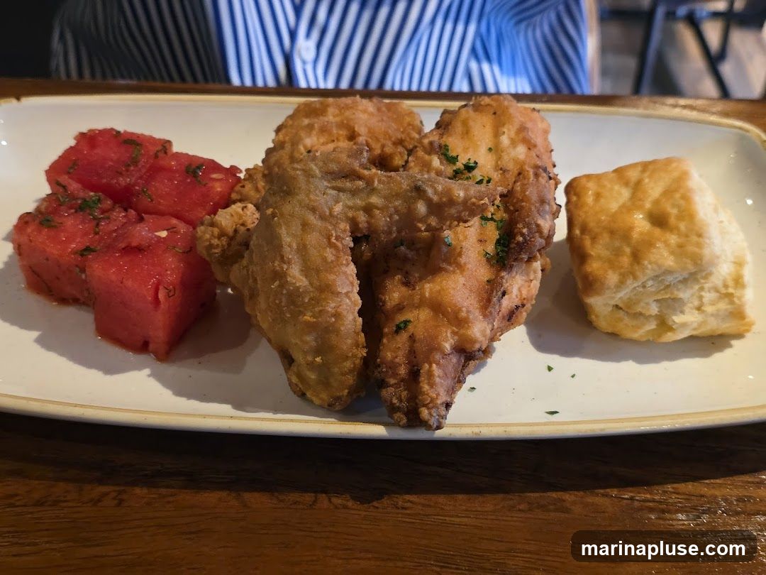 A plate of fried chicken, watermelon salad, and a biscuit sits on a wooden table. The chicken is golden brown and sprinkled with parsley. The watermelon is cubed and appears to be dressed with herbs. A flaky biscuit is to the right of the chicken. A person wearing a striped shirt is visible in the background.