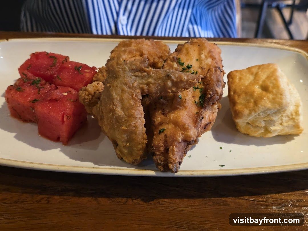 A plate of fried chicken, watermelon salad, and a biscuit sits on a wooden table. The chicken is golden brown and sprinkled with parsley. The watermelon is cubed and appears to be dressed with herbs. A flaky biscuit is to the right of the chicken. A person wearing a striped shirt is visible in the background.