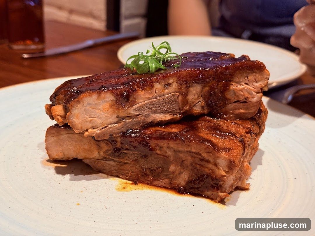 A close-up shot of a stack of barbecued ribs glazed with a dark, shiny sauce. The ribs are garnished with a few green scallions on top. The dish is served on a light-colored plate with a textured rim. In the background, a glass of dark liquid and a knife are visible on a wooden table.