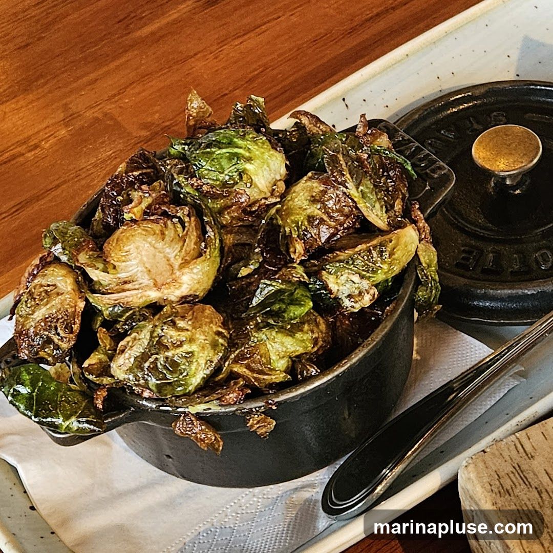 A close-up shot of roasted Brussels sprouts served in a small black cast-iron pot with a lid. The sprouts are crispy and glazed, with some leaves appearing caramelized. The pot is placed on a white textured napkin, which is on a rectangular white plate. A fork rests on the plate to the right of the pot. The background is a warm wooden surface.