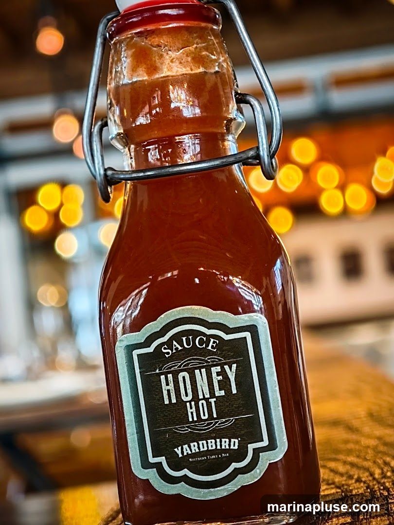 A close-up shot of a glass bottle filled with honey hot sauce. The bottle has a swing-top lid and a label that reads "SAUCE HONEY HOT YARDBIRD SOUTHERN TABLE + BAR". The background is blurred with warm, bokeh lights.