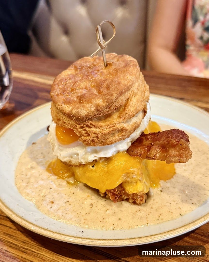 A close-up shot of a biscuit sandwich on a plate. The sandwich is stacked with a biscuit, fried chicken, melted cheese, a fried egg, and bacon. It is covered in gravy.