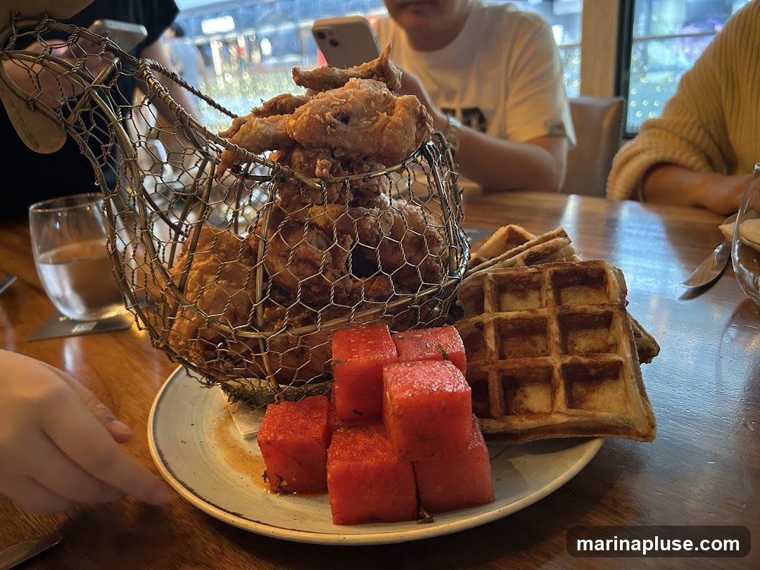 A plate of fried chicken served in a wire basket, accompanied by waffles and cubed watermelon. People are seated at the table in the background.