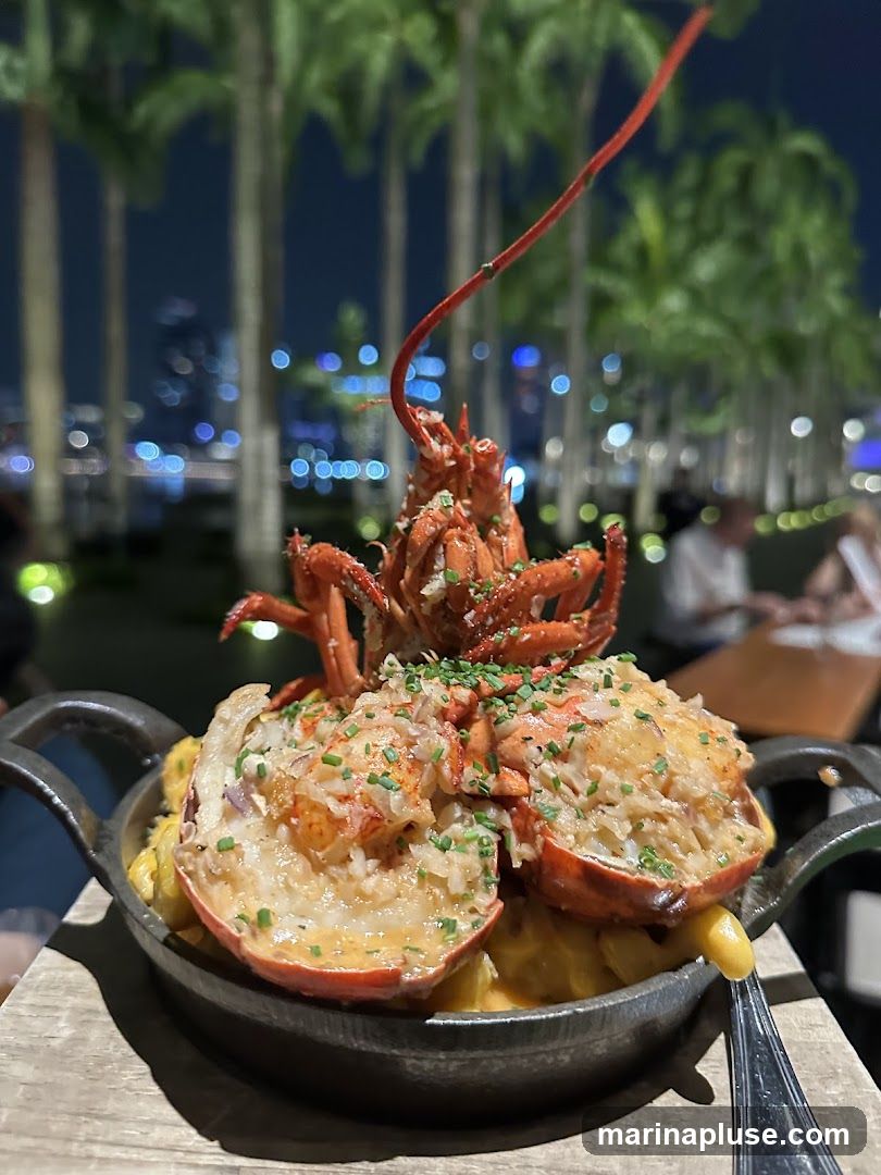 A close-up shot of a dish featuring lobster mac and cheese, served in a cast-iron skillet. The lobster is halved and topped with herbs and what appears to be garlic butter. The background shows a blurred view of palm trees and city lights at night, suggesting an outdoor dining setting.