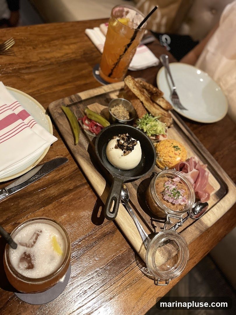 A wooden table with a charcuterie board featuring various dips, meats, pickles, and bread. Two cocktails are also visible on the table.