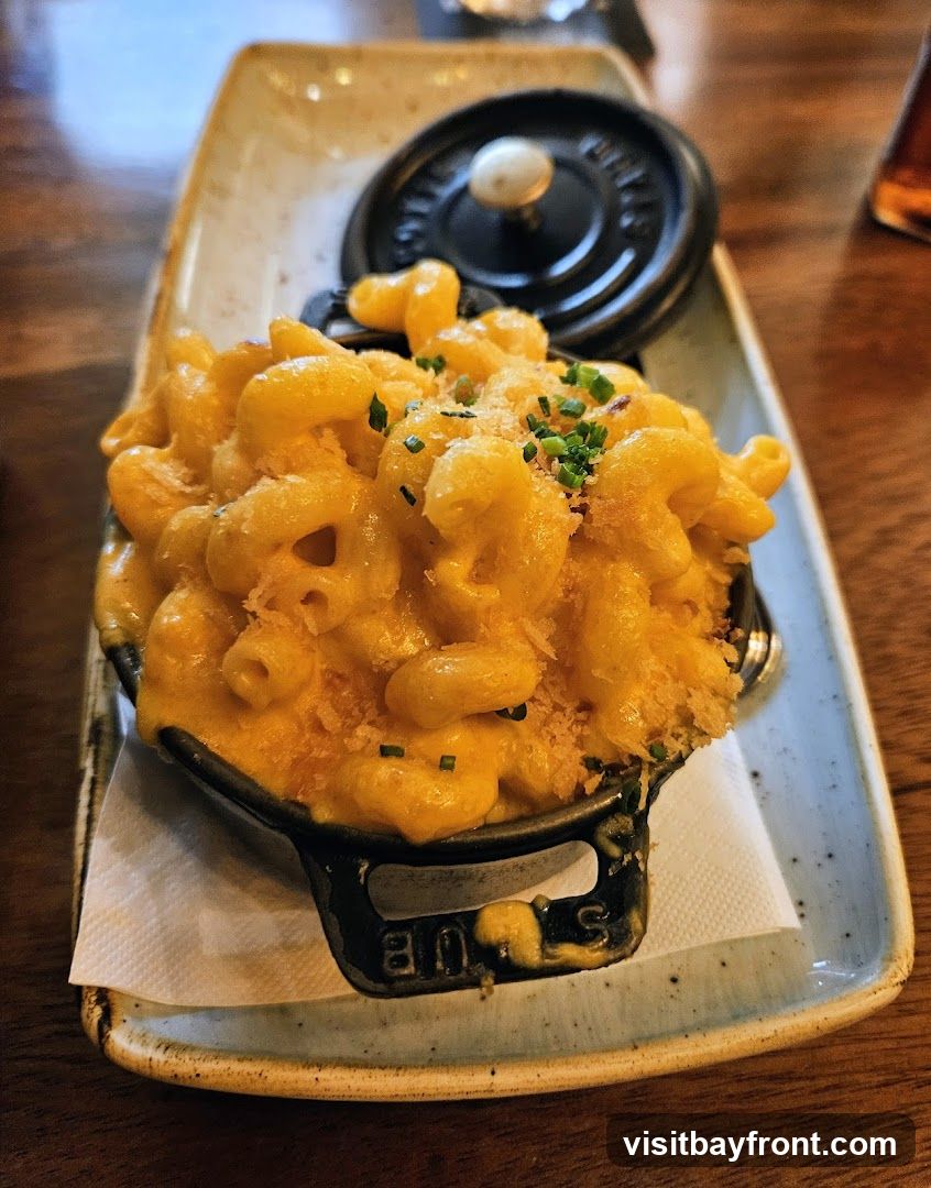 A serving of macaroni and cheese in a small black pot with a lid, garnished with chives and breadcrumbs. The pot is placed on a rectangular ceramic dish with a light brown glaze. The dish is on a wooden table.