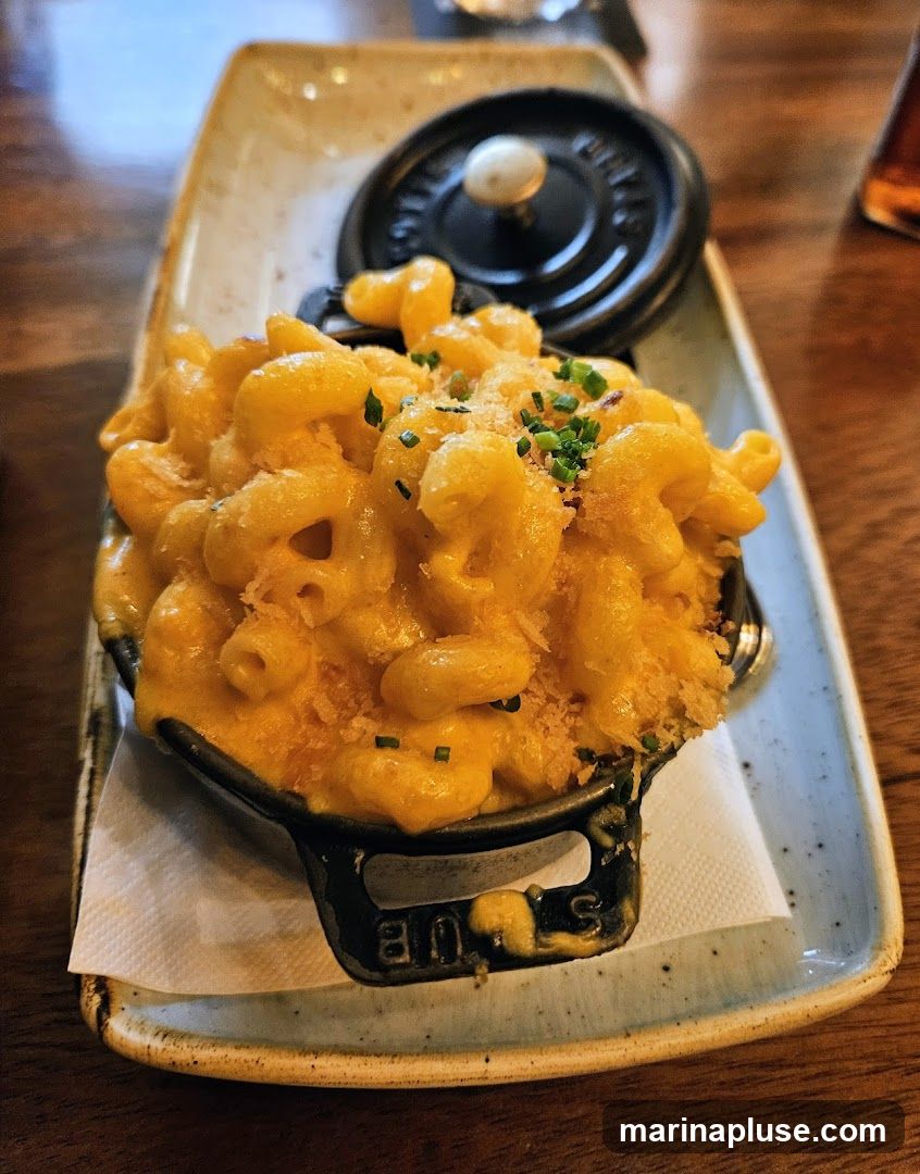 A serving of macaroni and cheese in a small black pot with a lid, garnished with chives and breadcrumbs. The pot is placed on a rectangular ceramic dish with a light brown glaze. The dish is on a wooden table.