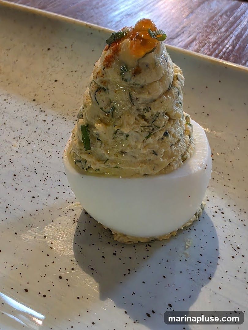 A close-up shot of a deviled egg appetizer. The egg white is filled with a creamy mixture, topped with what appears to be roe and chives, and dusted with paprika. The dish is served on a speckled white plate.