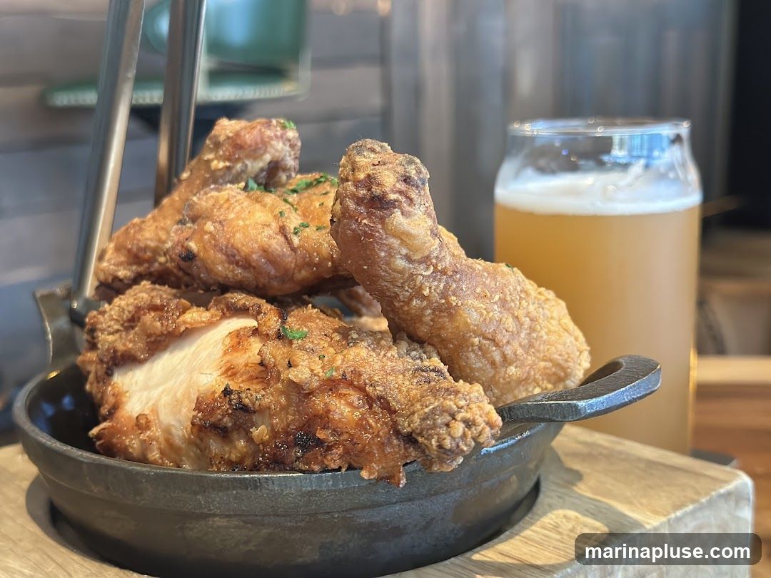 A close-up shot of a serving of crispy fried chicken pieces in a dark metal bowl, garnished with chopped herbs. To the right, a glass of light-colored beer with a frothy head is visible. The background is blurred, suggesting an indoor setting like a restaurant or bar.