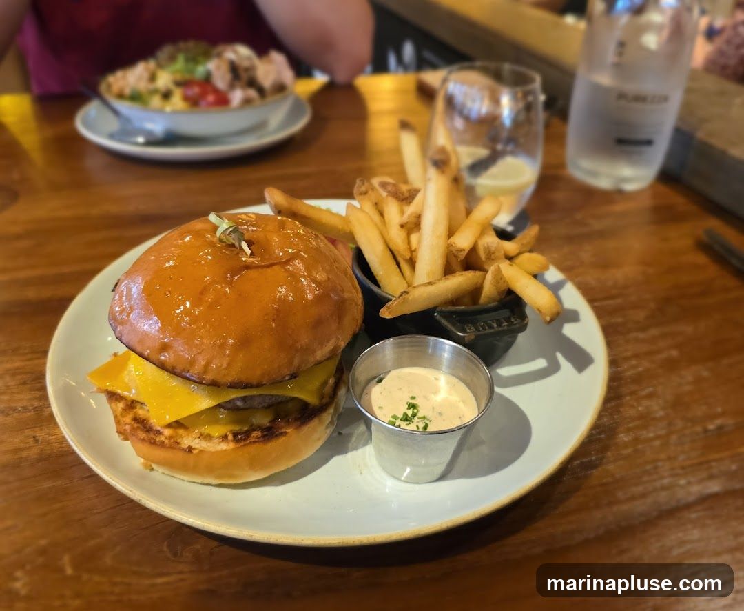 A close-up shot of a burger and fries served on a plate on a wooden table. The burger has a glossy bun, melted cheese, and a patty. A small metal cup of dipping sauce and a black Staub dish filled with french fries are next to the burger. In the background, a salad and a bottle of water are visible.
