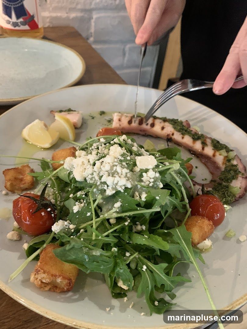 A close-up shot of a grilled octopus tentacle served with a salad of arugula, cherry tomatoes, feta cheese, and croutons. The dish is garnished with lemon wedges and a green sauce. A person's hands are visible, using a knife and fork to cut into the octopus.