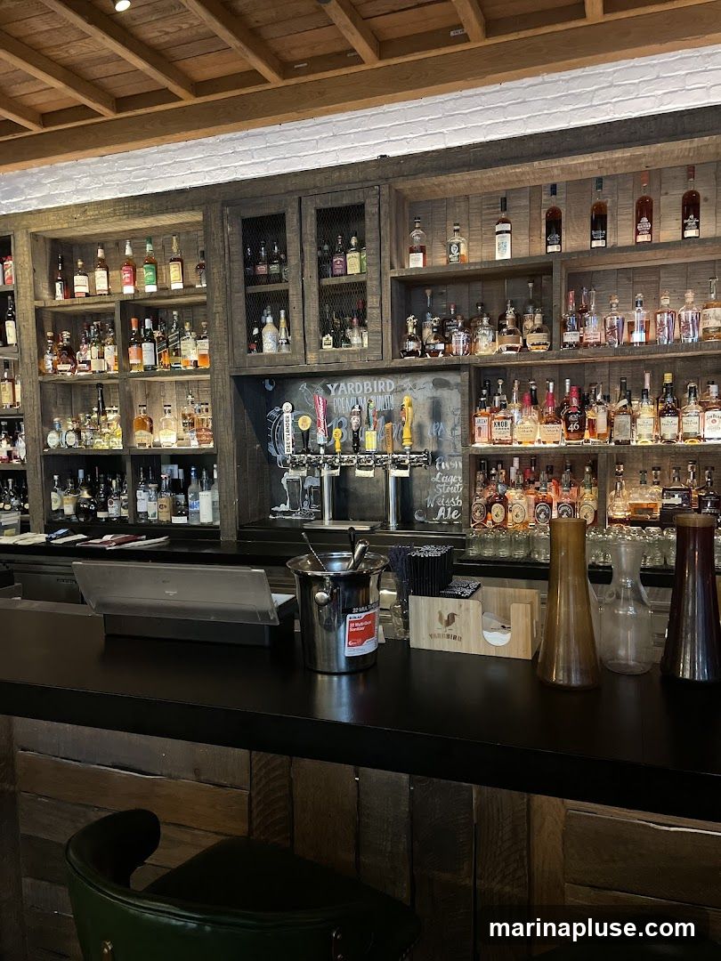 A well-stocked bar interior featuring numerous shelves filled with various liquor bottles. In the center, a row of beer taps is visible, with a chalkboard sign above them that reads "YARDBIRD PREMIUM LIQUOR". The bar counter is black, with a metal ice bucket, straws, and decorative glass vases. A green bar stool is partially visible in the foreground. The ceiling has exposed wooden beams.
