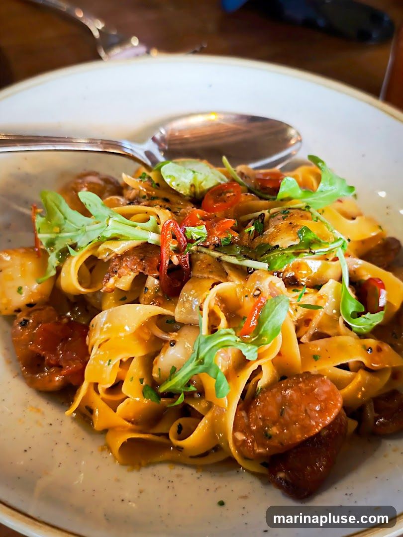 A close-up shot of a plate of fettuccine pasta with sausage, cherry tomatoes, and arugula. A spoon rests on the side of the plate.