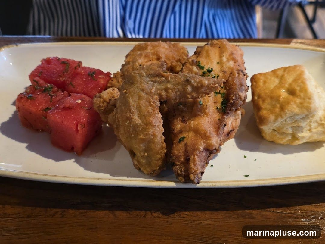 A plate of fried chicken pieces, cubed watermelon with herbs, and a biscuit is presented on a white platter. The platter is on a wooden surface. A person wearing a blue and white striped shirt is visible in the background.