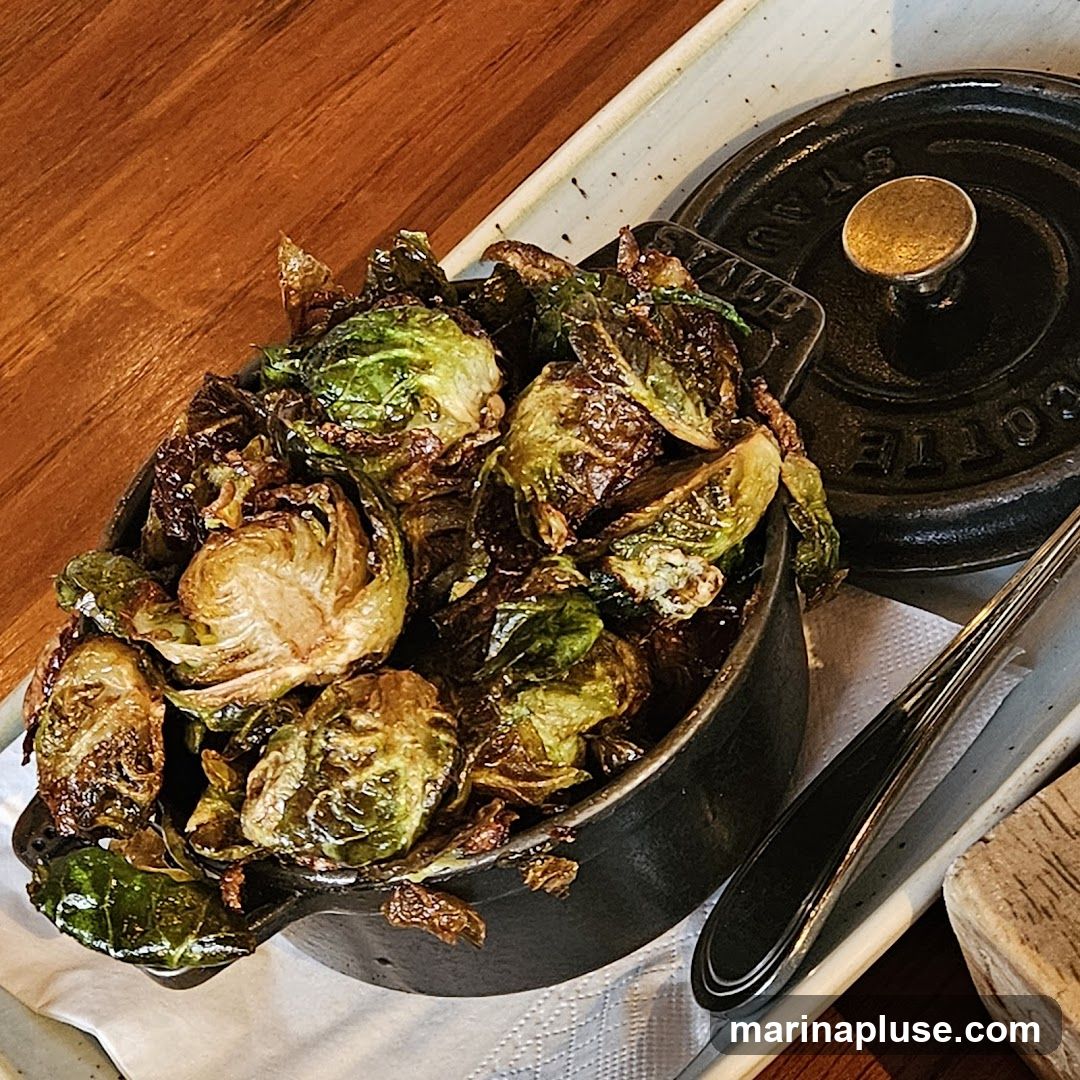 A close-up shot of a serving of crispy Brussels sprouts in a small black cast-iron pot. The sprouts are roasted to a deep brown and green, with some leaves appearing charred and crispy. The pot has a lid with a gold knob, and the brand name "STAUB" is visible on the lid. The dish is served on a rectangular white plate with a textured edge, placed on a wooden surface. A fork rests to the right of the pot.