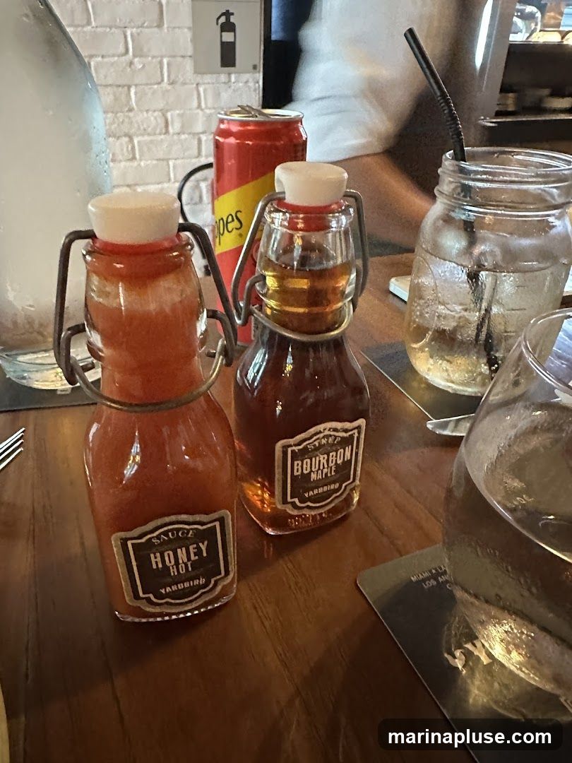 Two small glass bottles with flip-top lids are on a wooden table. The bottle on the left contains a red sauce labeled "SAUCE HONEY HOT YARDBIRD". The bottle on the right contains a dark amber liquid labeled "SYRUP BOURBON MAPLE YARDBIRD". A red can with yellow lettering is behind the bottles. A glass jar with a straw and a glass with ice and a straw are also visible. A white brick wall is in the background.