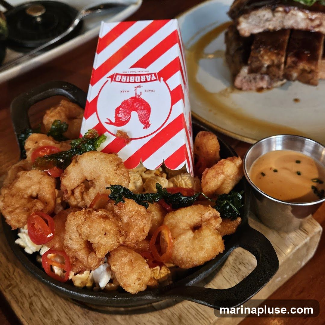 A close-up shot of a dish featuring fried shrimp served in a black cast-iron pan. The shrimp are golden brown and mixed with some greens and red pepper slices. A small red and white striped box with a logo is placed in the center of the dish. To the right, a small metal cup holds a creamy orange dipping sauce. In the background, a plate of sliced ribs is visible.