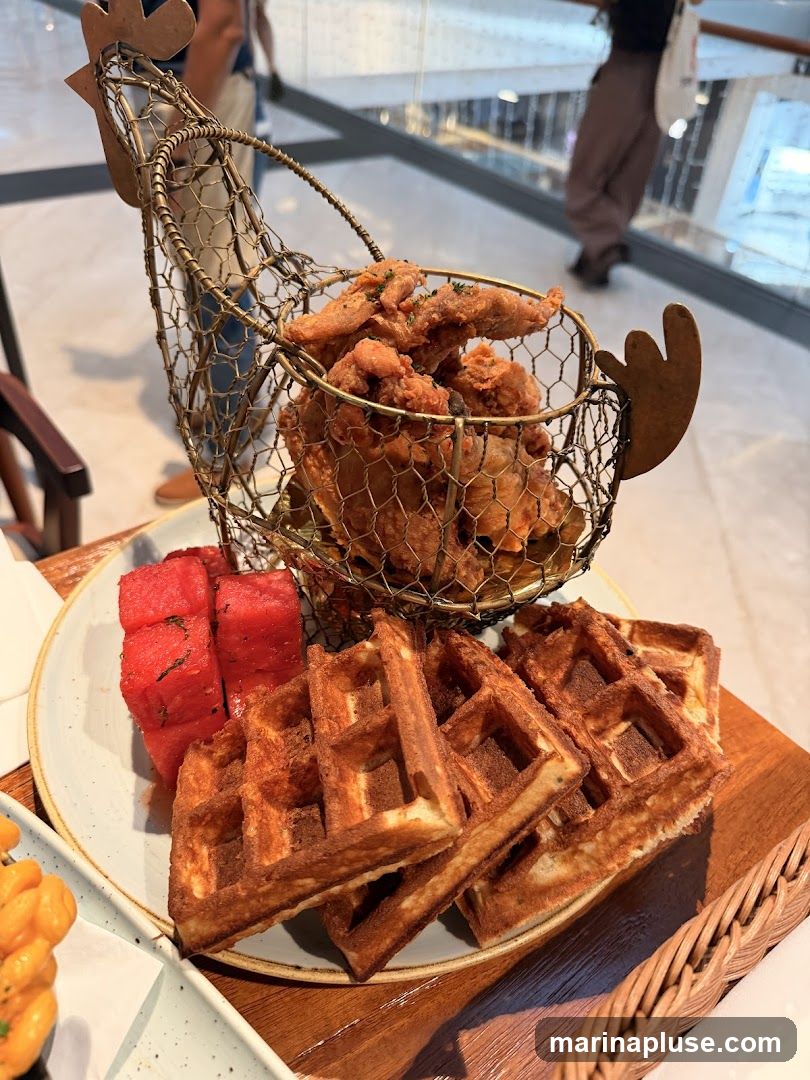 A plate of fried chicken served in a wire chicken-shaped basket, accompanied by waffles and watermelon slices. The food is presented on a wooden board. In the background, blurred figures are visible in what appears to be a mall or public space.