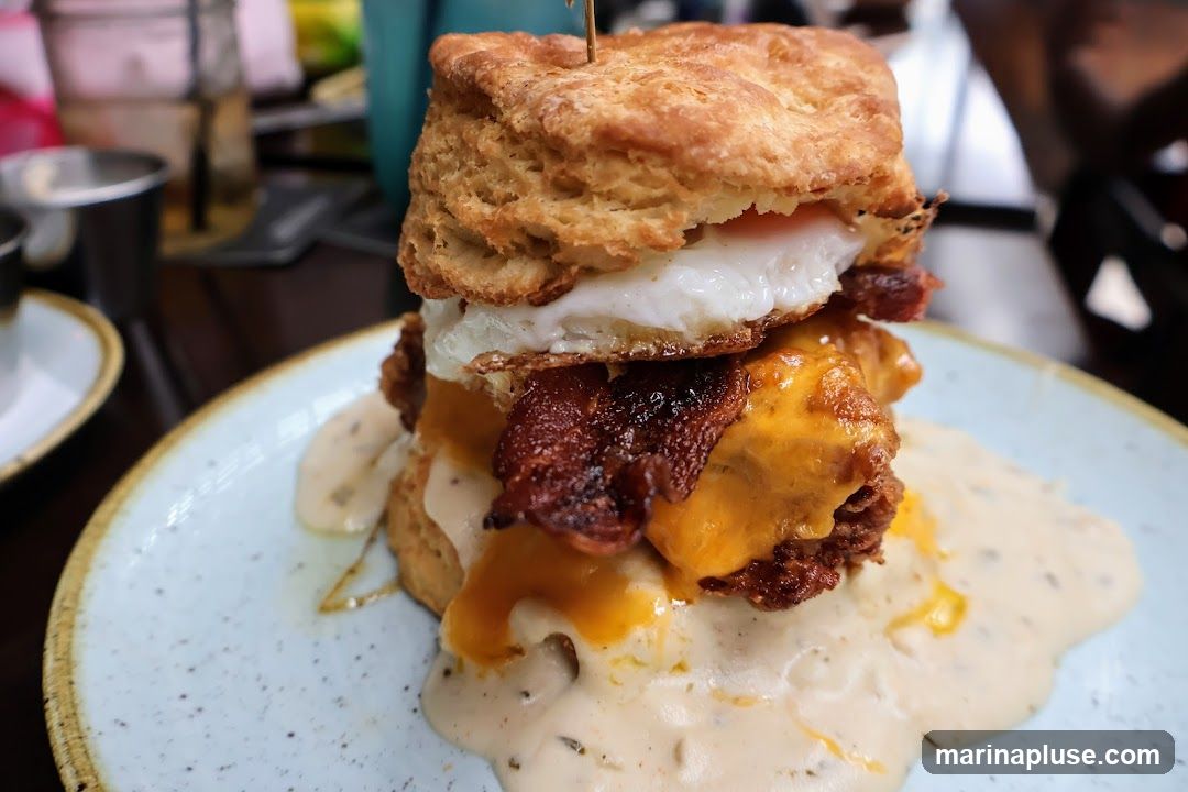 A tall biscuit sandwich is served on a speckled blue plate, generously covered in gravy. The sandwich is layered with a thick biscuit, a fried egg, crispy bacon, melted cheese, and what appears to be fried chicken. The gravy is creamy and has visible herbs or spices. In the background, out of focus, are other dining elements like metal cups and a blue object.