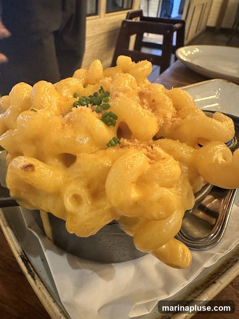 A close-up shot of a serving of macaroni and cheese, topped with chopped chives and breadcrumbs. The pasta is coated in a thick, creamy cheese sauce. The dish is served in a dark, round dish, which sits on a light-colored plate with a textured rim. In the background, blurred elements of a restaurant interior are visible, including wooden furniture and a person's shoulder.
