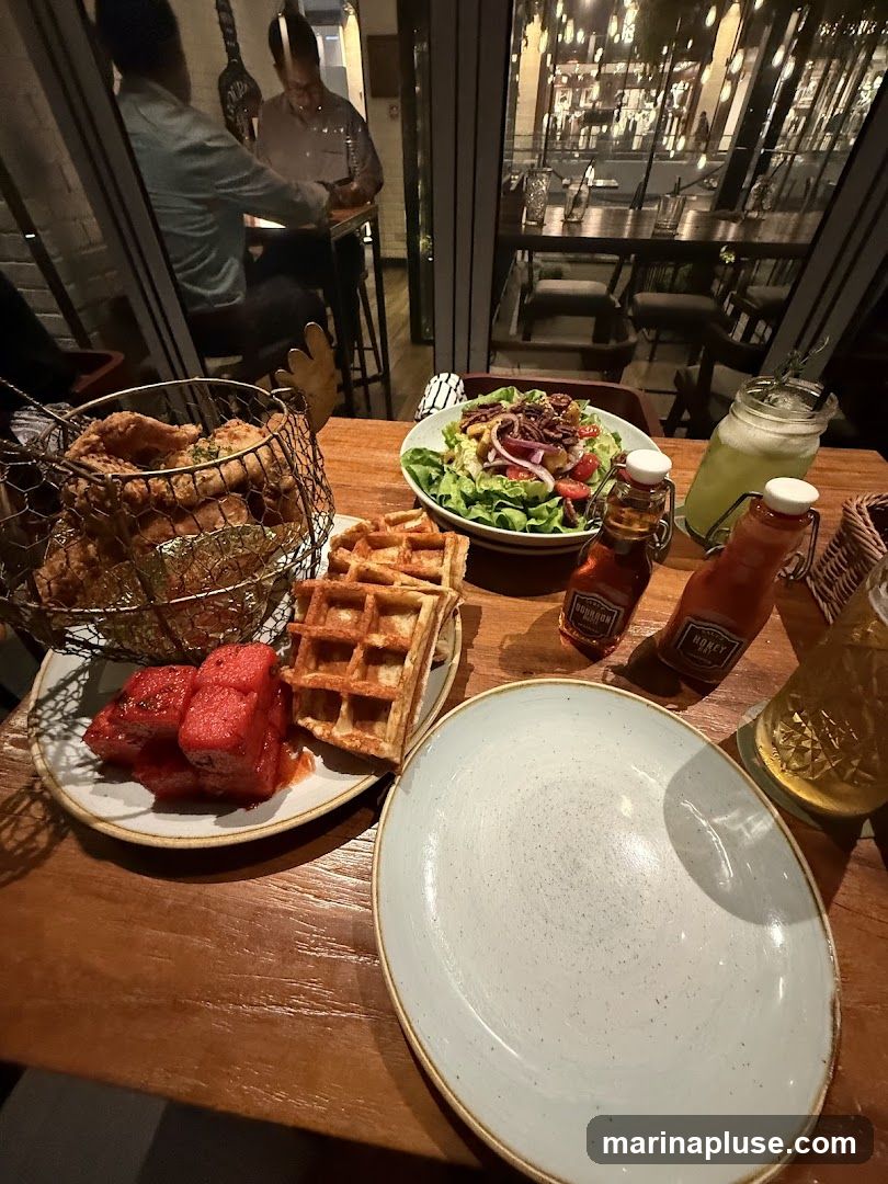 A wooden table is set with a meal, including fried chicken in a wire basket, waffles, watermelon slices, and a salad. Two bottles labeled 'Bourbon' and 'Honey' are on the table, along with a glass of green liquid and another drink. In the background, through a large window, the interior of a restaurant with tables and lights is visible, and two men are seated at a high table.