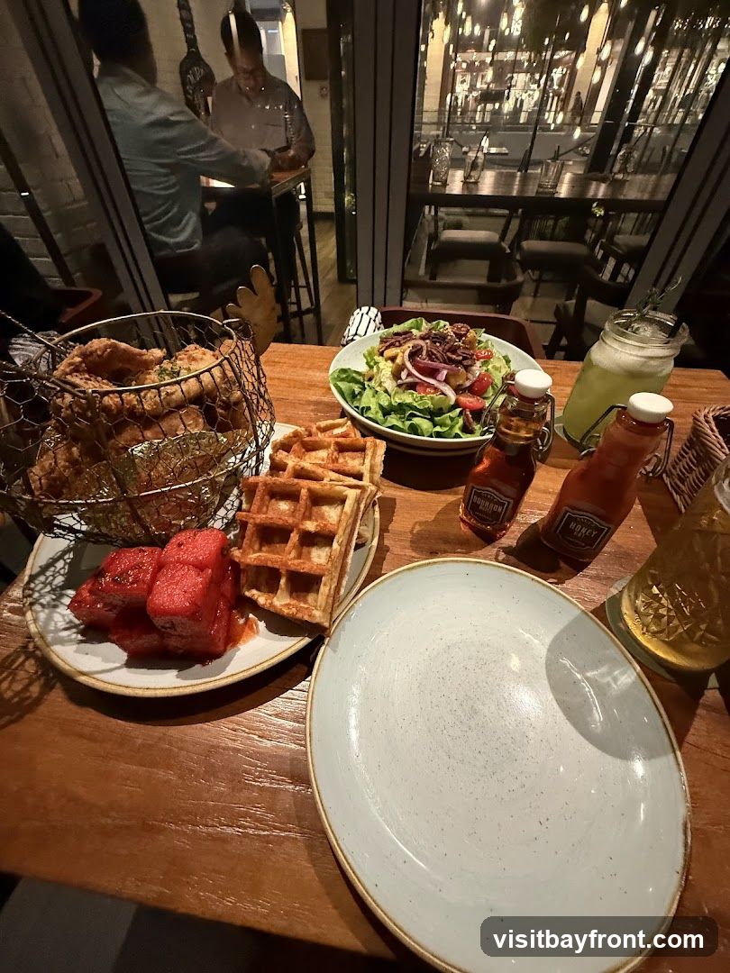 A wooden table is set with a meal, including fried chicken in a wire basket, waffles, watermelon slices, and a salad. Two bottles labeled 'Bourbon' and 'Honey' are on the table, along with a glass of green liquid and another drink. In the background, through a large window, the interior of a restaurant with tables and lights is visible, and two men are seated at a high table.