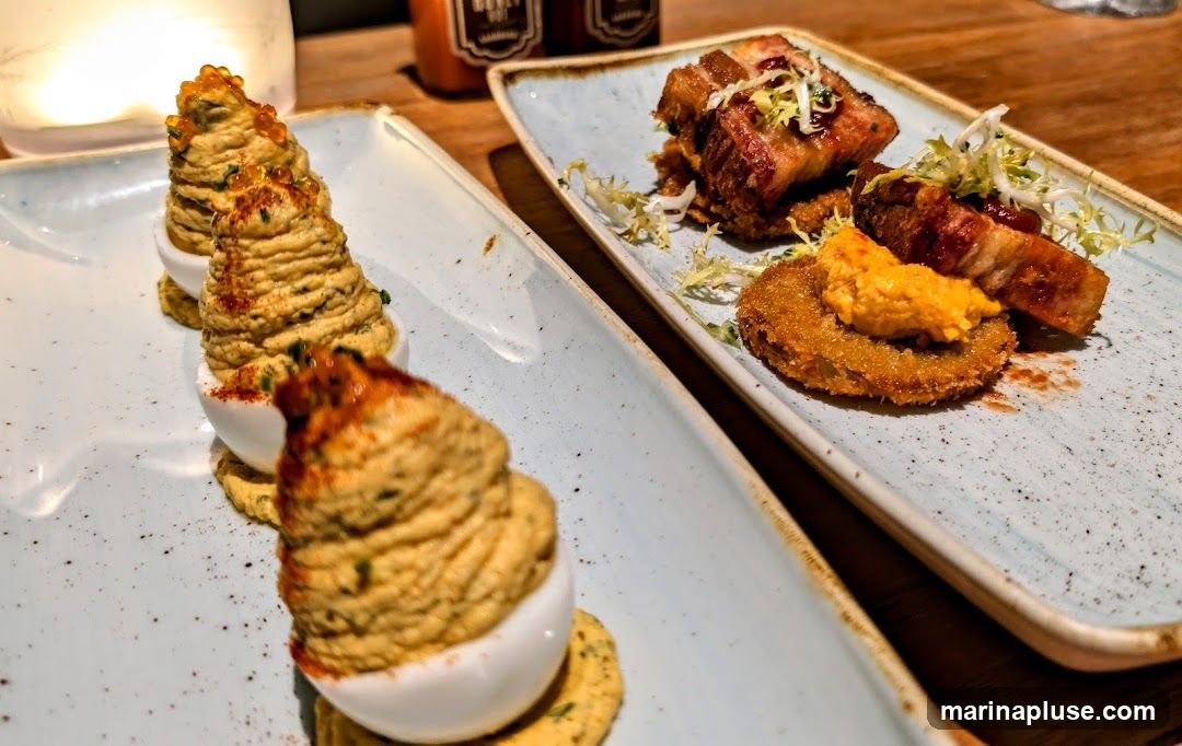 Two plates of appetizers are shown. The plate in the foreground contains several deviled eggs topped with a creamy filling and red caviar. The plate in the background features pieces of pork belly served with a fried green tomato and a dollop of orange sauce. Two bottles of hot sauce are visible in the background.