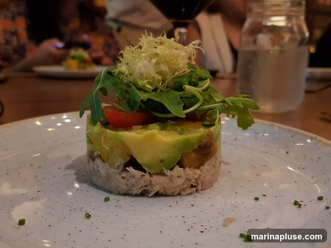 A close-up shot of a layered appetizer on a white speckled plate. The appetizer is a tower with a base of shredded tuna, followed by layers of avocado and a cherry tomato, topped with frisée lettuce and arugula. In the background, out of focus, are diners, a glass of red wine, and a mason jar filled with water.