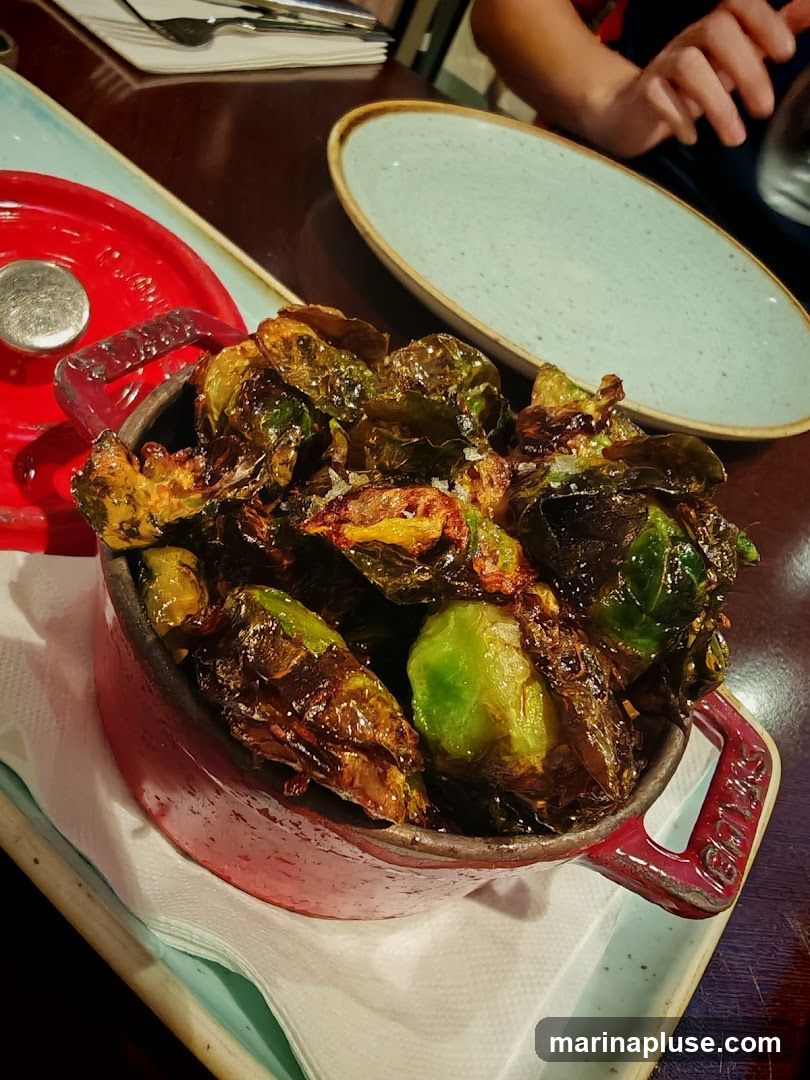 A close-up shot of roasted Brussels sprouts served in a red pot. The sprouts are glistening and appear crispy, with some leaves slightly charred. The dish is placed on a white napkin, which is on a light blue serving platter. In the background, there are plates and cutlery, suggesting a dining setting.