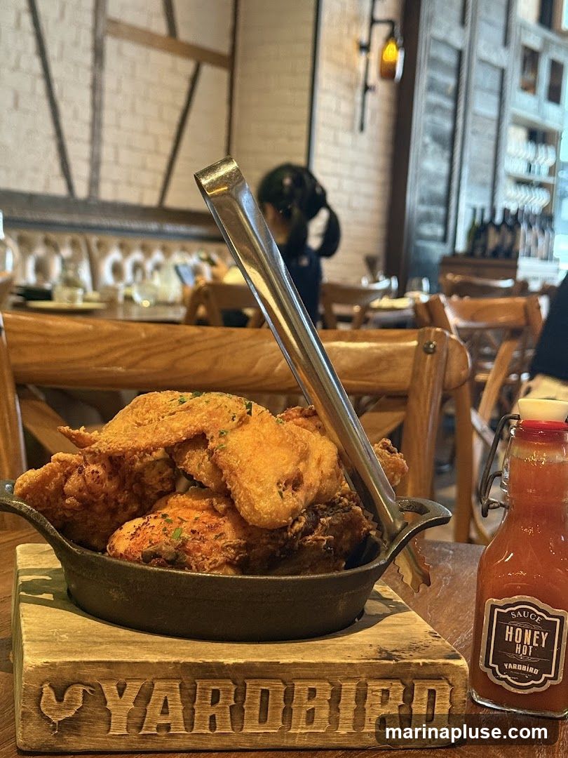 A close-up shot of a cast-iron skillet filled with golden-brown fried chicken pieces, served on a wooden board with the word "YARDBIRD" carved into it. Tongs are placed in the chicken. To the right, a small glass bottle of "HONEY HOT SAUCE" from YARDBIRD is visible. The background shows a restaurant interior with wooden furniture and patrons dining.