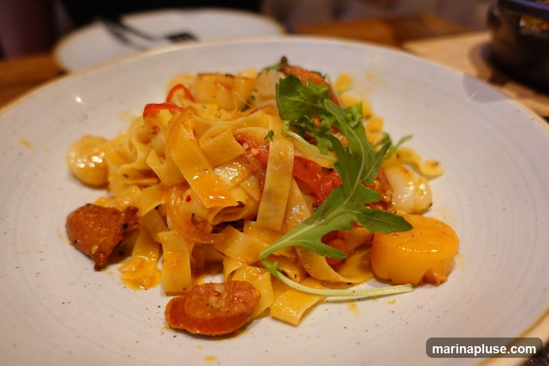 A close-up shot of a plate of pasta with scallops and sliced sausage. The pasta is wide and flat, coated in a reddish-orange sauce with visible pieces of tomato and chili flakes. Several seared scallops are scattered throughout the dish, along with slices of cooked sausage. A sprig of arugula garnishes the top. The plate has a speckled, off-white finish with a light brown rim. In the background, out of focus, are other plates and a dark object, suggesting a restaurant setting.