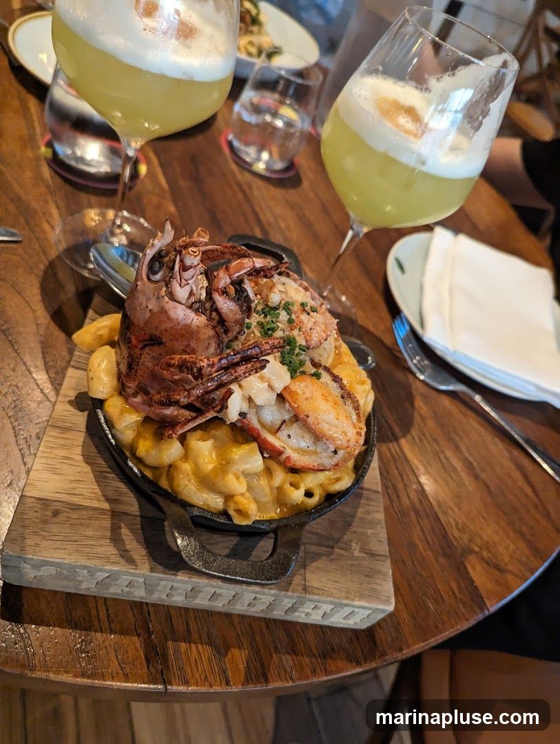 A close-up view of a serving of lobster mac and cheese, topped with a whole lobster claw and tail, served in a cast-iron skillet on a wooden board. Two glasses of yellow cocktails with foam are visible in the background, along with water glasses and napkins. The setting appears to be a restaurant table.