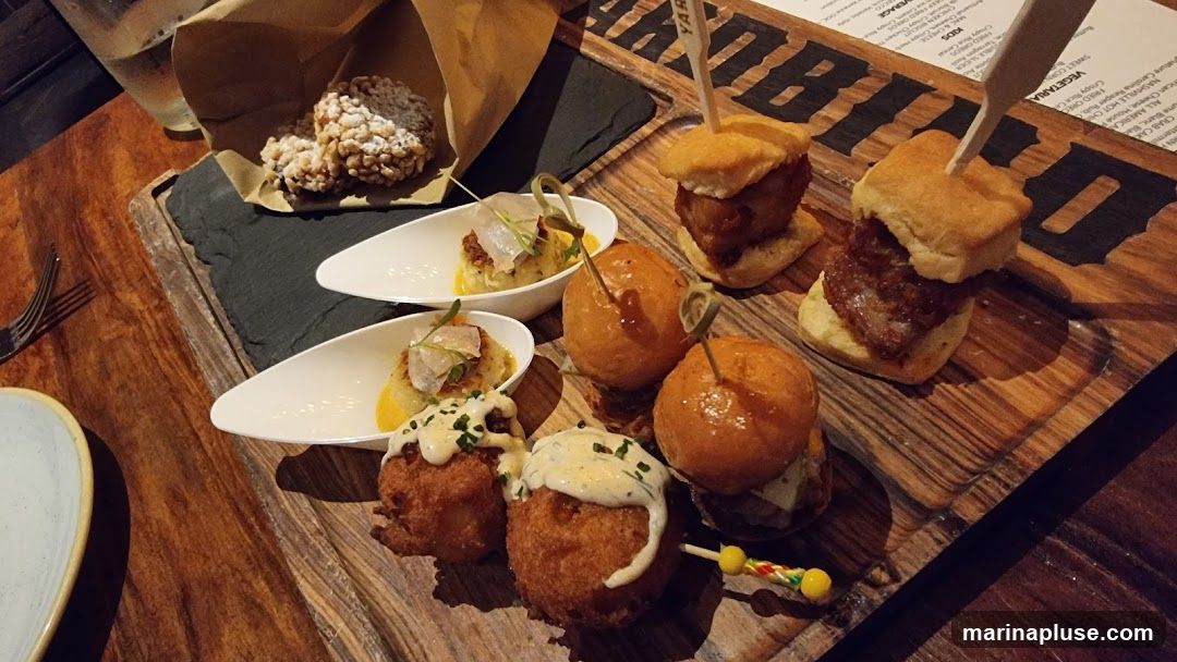 A wooden board with various appetizers, including sliders, crab cakes, and fried items. A glass of water and a fork are also visible.