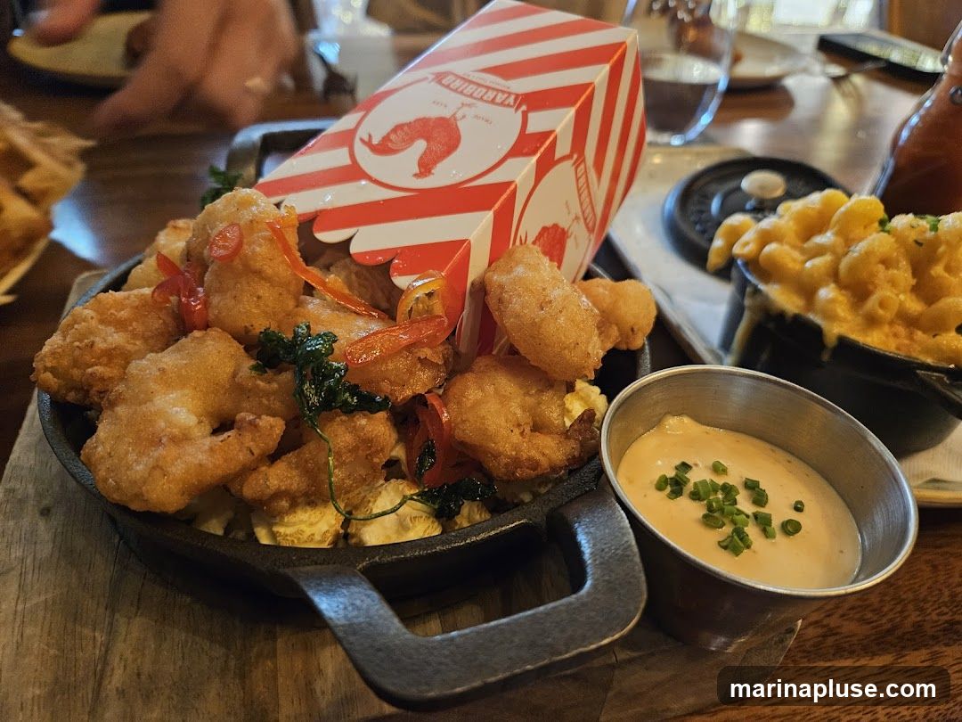 A close-up shot of a platter of fried food, possibly chicken tenders or shrimp, served in a cast-iron skillet. The food is garnished with red pepper slices and herbs. A red and white striped box with a rooster logo is placed on top of the fried food. To the right, a small black pot contains macaroni and cheese, and a metal ramekin holds a dipping sauce topped with chives. The meal is presented on a wooden surface, with other diners and a bottle of sauce visible in the blurred background.