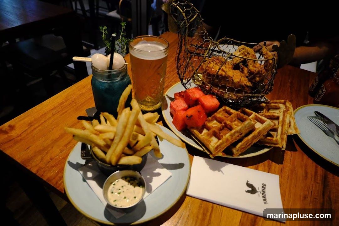 A meal consisting of fried chicken served in a wire basket shaped like a chicken, waffles, watermelon slices, french fries, a blue cocktail with lychee, and a glass of beer on a wooden table. A napkin with the logo "YARDBIRD" is also visible.