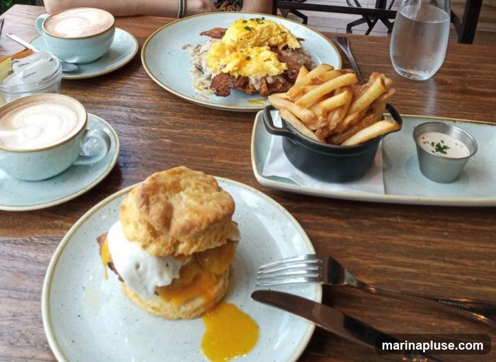 A wooden table is set with a brunch meal. In the foreground, a biscuit sandwich with egg and cheese is served on a plate with a fork and knife. To the right, a small black pot holds a serving of french fries with a side of dipping sauce. In the background, another plate features scrambled eggs, bacon, and gravy over what appears to be a biscuit or bread. Two cups of latte art coffee are also visible on the table. A glass of water sits in the upper right corner.