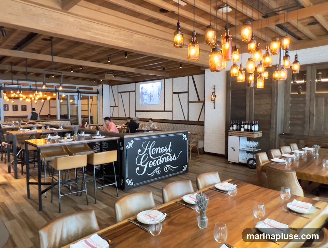 The interior of a restaurant with wooden tables and chairs, a bar area, and decorative lighting. A sign on the bar reads "Honest Goodness". There are patrons seated at tables and a wine display cart.