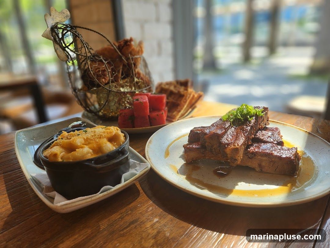 A close-up shot of a meal on a wooden table. In the foreground, a plate of braised short ribs topped with green onions sits in a pool of sauce. To the left, a small black ramekin holds macaroni and cheese, sprinkled with breadcrumbs. In the background, a wire chicken basket contains fried chicken pieces, and a small plate holds several cubes of watermelon and some waffles. The scene is lit by natural light coming from a window.